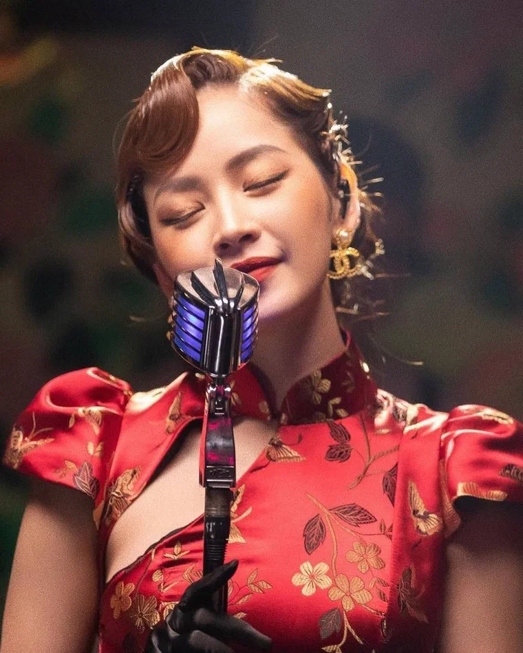 Chi Pu tóc nâu rẽ ngôi lệch, uốn sóng nước sát đầu kiểu thập niên 1930, nhắm mắt hát vào micro cổ, mặc sườn xám đỏ tay bồng