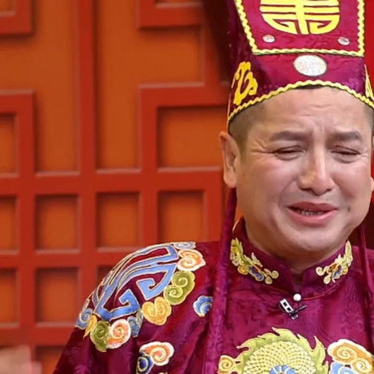 Chí Trung trong áo bào đỏ thẫm thêu hoa văn, đội mũ Táo đỏ cao có chữ Thọ vàng, nhăn mặt diễn trên sân khấu