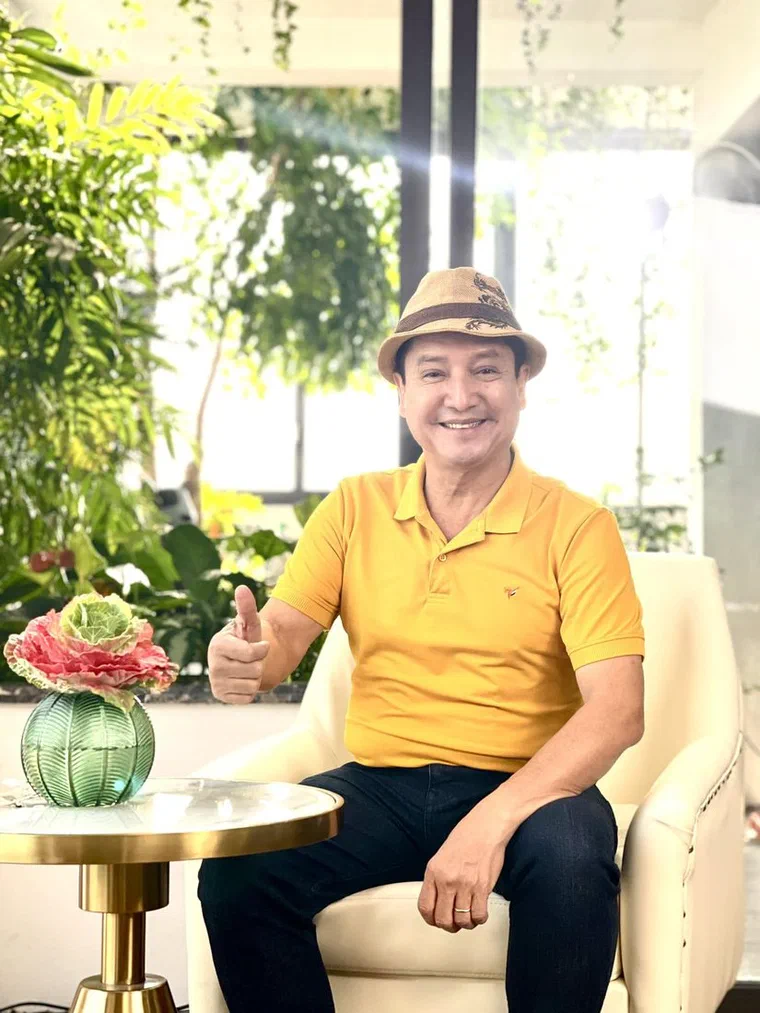 Chí Trung đội mũ phớt cói màu nâu nhạt viền đen, áo polo vàng, ngồi ghế bành giơ ngón cái, cười tươi