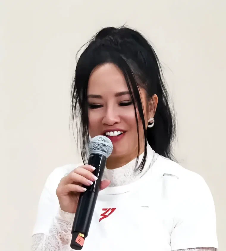 Hồng Nhung tóc đen buộc cao trên đỉnh đầu, nhiều lọn tóc mai dài buông hai bên má, đang cầm micro nói chuyện