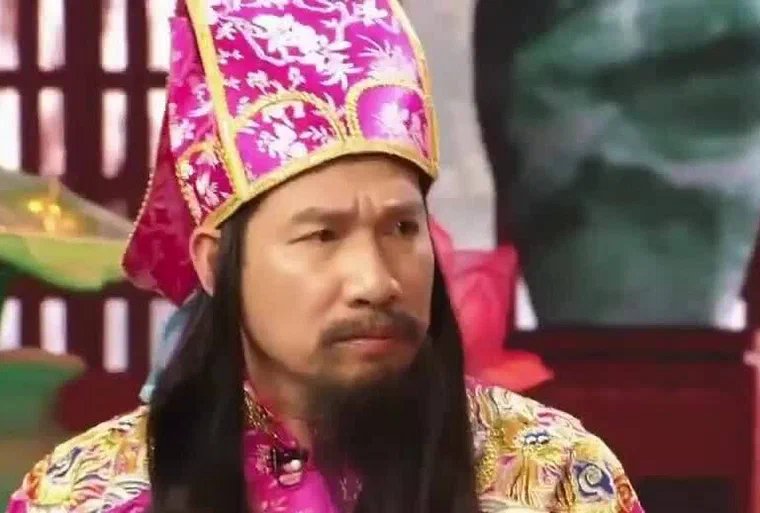 Quang Thắng trong Táo Quân 2022, đội mũ Táo hồng, tóc giả đen dài rủ quá vai, râu giả đen dài, áo bào hồng thêu