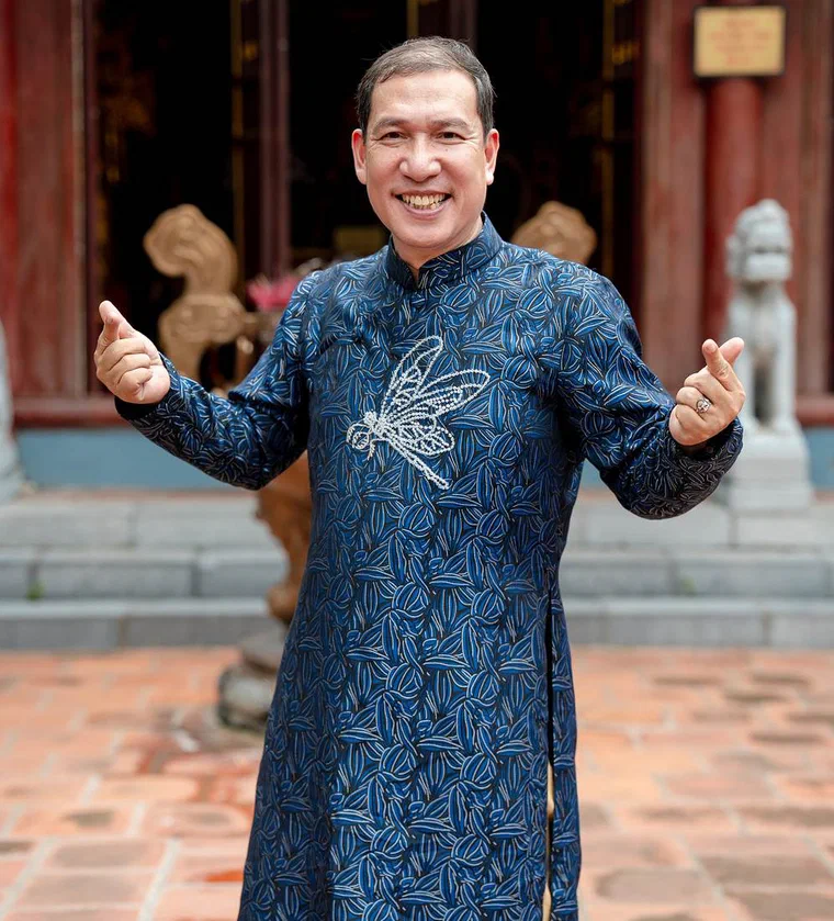Quang Thắng mặc áo dài xanh đậm thêu chuồn chuồn bạc, dang tay cười trước sân đình, tóc ngắn vuốt ngược, hai bên mai đã điểm bạc