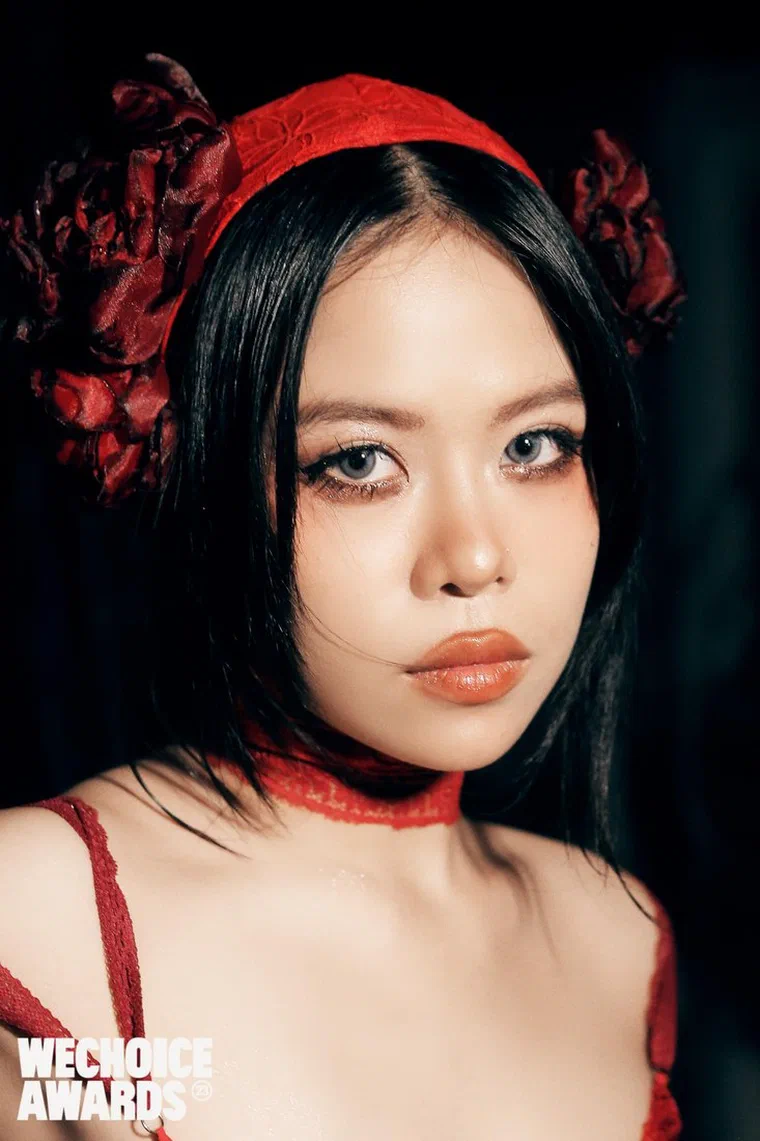tlinh tóc đen rẽ ngôi giữa ép sát vào da đầu, đội bờm đỏ gắn hai bông hoa vải, đeo choker ren đỏ
