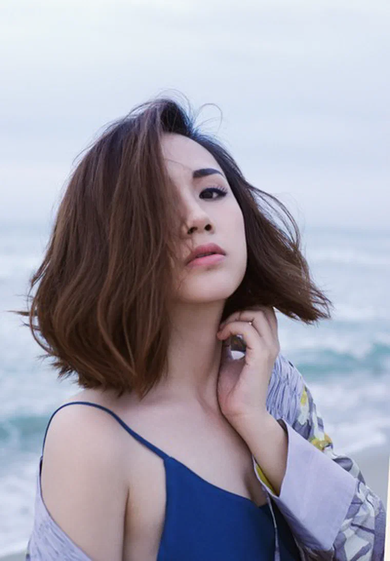 Trâm Nguyễn tóc bob nâu hạt dẻ dài chạm vai, rẽ ngôi lệch, gió thổi rối, nền biển