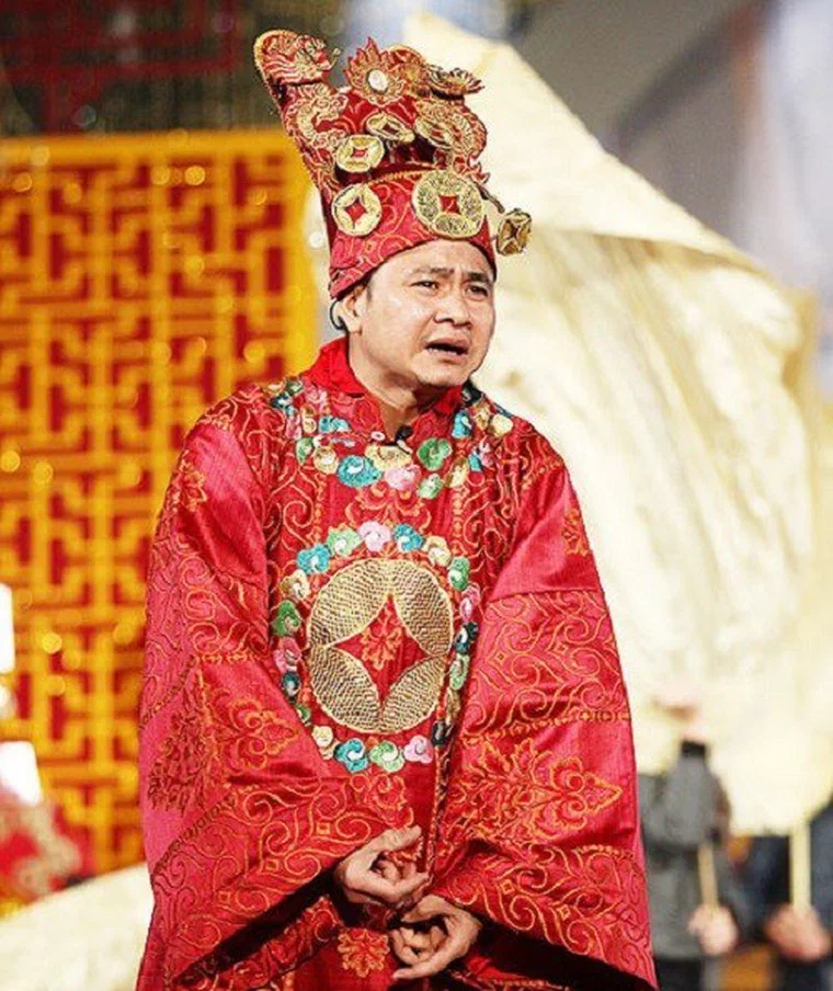 Tự Long trong áo bào đỏ thêu kín hoa văn, đội mũ Táo cao gắn đồng tiền vàng, chỉ lộ phần tóc mai đen tỉa sát trên vành tai
