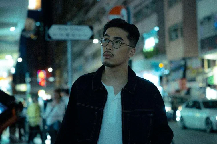 Vũ. đứng ngoài phố đêm, tóc đen vuốt ngược hết khỏi trán, hai bên tỉa sát ôm đầu, đeo kính gọng tròn