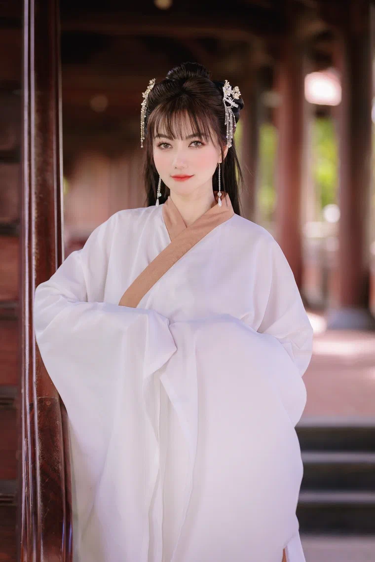 Yuni Boo với tóc đen dài, phần đỉnh vấn búi tròn cài hai cây trâm có tua ngọc, mái thưa ngang chân mày, mặc áo cổ trang trắng viền nâu